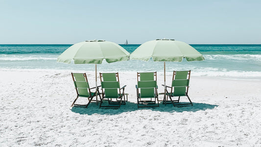 Four green beach chairs and two green umbrellas on a white, sandy beach next to the ocean