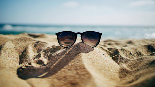 Pair of Ray Ban sunglasses sitting on a small pile of sand with the ocean in the background